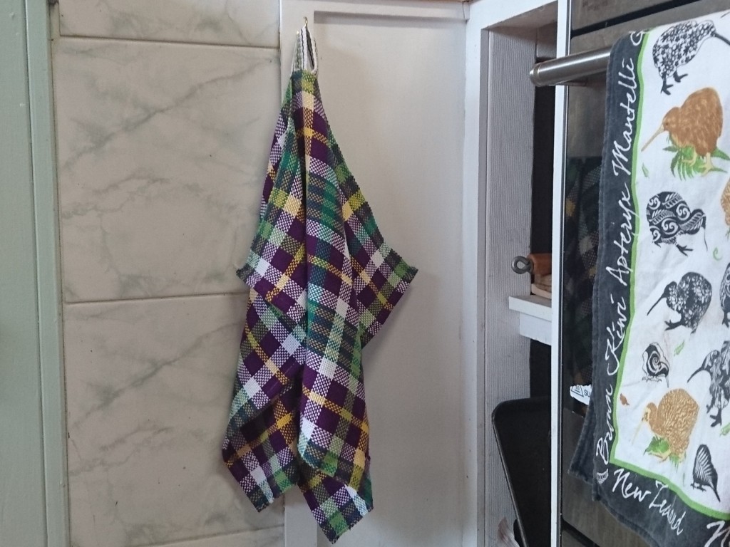 A plaid tea towel hanging from a hook in the corner of a kitchen, beside an oven, there is another tea towel hanging on the oven door with kiwi birds on it, and shelves with some pans and trays poking out.
